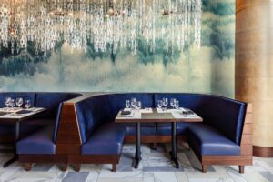 Del Frisco's Double Eagle Steakhouse San Diego spacious, private booths upholstered in plush navy blue leather. Brass lighting elements are surrounded by a glass-chain ceiling fixture. The walls are clad with textured, abstract imagery in blues and whites, reminiscent of the ocean.