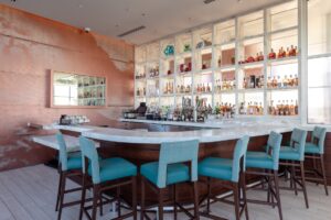Aqua upholstered chairs surround a gleaming white bar. Light coral walls are accented with white liquor shelves behind the bar in the Del Frisco's Double Eagle Steakhouse San Diego