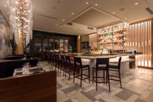 A nighttime photo of one of the bars at Del Frisco's Double Eagle steakhouse San Diego. Custom Light fixtures twinkle, and liquor shelves behind the bar are backlit with warm light